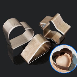 May include: Three metal craft punches in the shapes of a heart, star, and teardrop. The heart punch is shown in use, punching a heart shape into a piece of leather.