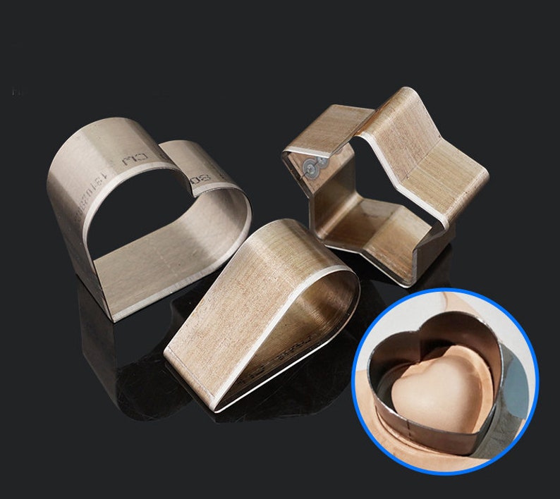 May include: Three metal craft punches in the shapes of a heart, star, and teardrop. The heart punch is shown in use, punching a heart shape into a piece of leather.