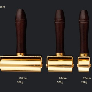 May include: Three gold-colored leather working tools with wooden handles. The tools are different sizes: 100mm, 60mm, and 26mm. The tools are on a black background.
