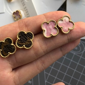 Può includere: Un paio di orecchini a forma di trifoglio dorati con inserti a motivi diversi. Un paio ha un inserto a motivi marroni e neri, l'altro paio ha un inserto a motivi rosa e bianchi.