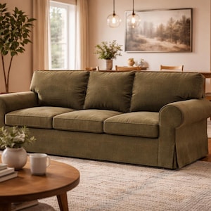 Op de afbeelding: Een olijfgroene driezitsbank met gerolde armen in een woonkamer. Een ronde houten salontafel staat voor de bank. Links van de bank staat een houten bijzettafel met een lamp. Onder de salontafel ligt een vloerkleed.