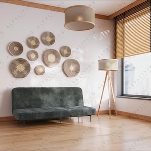 Puede incluir: Un sofá futón gris oscuro con un marco de madera se encuentra en una habitación con una pared blanca y un suelo de madera. La pared está decorada con una colección de decoración de pared redonda tejida en tonos marrones y beige. Una lámpara con una pantalla beige cuelga del techo. Una lámpara de pie con una pantalla blanca y una base de trípode se encuentra en la esquina de la habitación. Una ventana con persianas de madera está abierta, revelando una vista del horizonte de la ciudad.