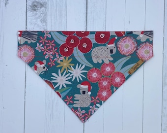 May include: A teal dog bandana with a festive floral and animal print. The design features red flowers, koalas, and mice wearing Santa hats. The bandana is triangular in shape and has a small tag with text on the right side.