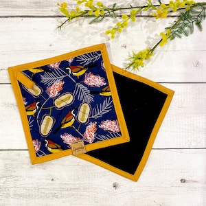 May include: Two square pot holders. One has a navy blue bird and floral pattern, edged with gold trim. The other is plain black with gold trim. A small tag with text is attached to the patterned pot holder.