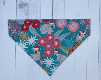 May include: A teal dog bandana with a repeating pattern of koalas and flowers. The koalas are wearing Santa hats, and the flowers are red, pink, and white. The bandana is made of fabric and has a triangular shape.