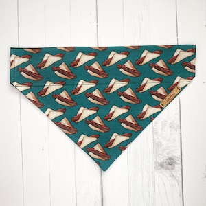 May include: Teal blue dog bandana featuring a repeated pattern of illustrated hot dogs in white bread. The bandana has a brown leather tag with the word "Sparky" printed on it.