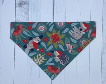 May include: Teal dog bandana with a repeating pattern of Australian animals, including a koala, echidna, and lizard, wearing Santa hats. The design also features red and white flowers and green leaves.