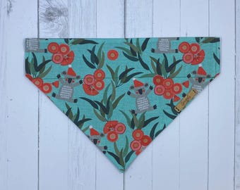 May include: A turquoise pet bandana with a repeating pattern of gray koalas wearing Santa hats, red flowers, and green leaves. The bandana is triangular in shape and made of fabric.