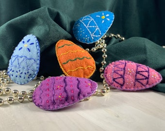 May include: Five felt Easter eggs with embroidered designs. The eggs are blue, orange, purple, and pink with white embroidery. The eggs are on a white surface with a silver beaded garland.