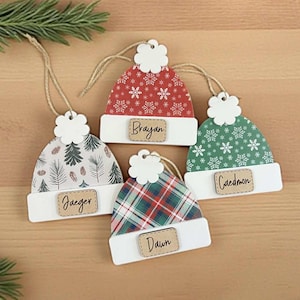 May include: Four wooden Christmas ornaments shaped like winter hats. Each hat features a different pattern: snowflakes, pine trees, and plaid. Each ornament has a white brim and a small wooden tag with a name.