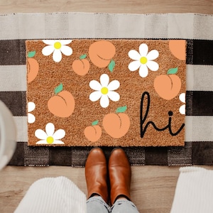 Puede incluir: Una alfombra de puerta marrón con un diseño de melocotones y margaritas, y la palabra "hi" en escritura negra. La alfombra está colocada sobre una alfombra a cuadros en blanco y negro. Se ven botas marrones.