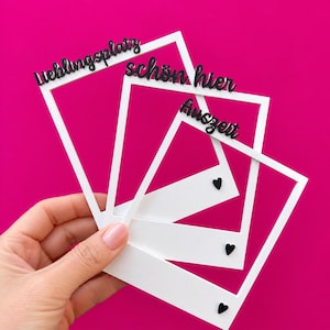 Könnte beinhalten: Drei weiße, rechteckige Fotorahmen, die fächerförmig angeordnet sind. Jeder Rahmen trägt oben einen schwarzen, kursiven Schriftzug mit den Wörtern "Lieblingsplatz", "schön hier" und "Auszeit". Kleine schwarze Herzsymbole zieren die unteren Ränder.