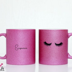 Könnte beinhalten: Zwei rosa Glitzerbecher mit schwarzen Designs. Ein Becher hat den Namen "Suzanne" darauf geschrieben. Der andere Becher hat ein Paar geschlossene Augen mit Wimpern.