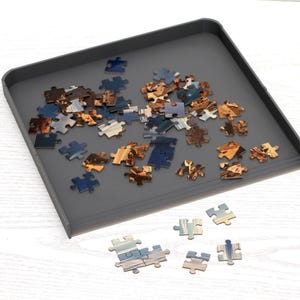 Könnte beinhalten: Eine graue Puzzleschale mit verschiedenen Puzzleteilen. Die Teile sind in Blau-, Braun- und Goldtönen gehalten. Mehrere Teile sind außerhalb der Schale auf einer weißen Holzoberfläche verstreut. Die Schale hat einen erhöhten Rand.