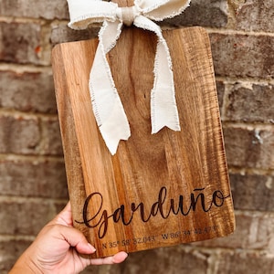 May include: A rectangular wooden chopping board with a handle and a cream ribbon tied at the top. The word "Garduno" is inscribed in cursive, along with coordinates. The board has a natural wood grain.