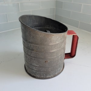 Pode incluir: Um peneira de farinha vintage de metal com uma alça vermelha. A peneira tem marcas para medir xícaras.
