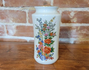 May include: White ceramic vase with a floral design featuring orange, pink, blue, and yellow flowers. The vase has a gold rim.