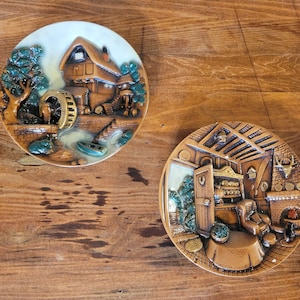 Może przedstawiać: Dwa dekoracyjne talerze ceramiczne z szczegółowymi scenami reliefowymi. Jeden przedstawia młyn wodny i domek, drugi przytulne wnętrze z kominkiem i fotelem. Talerze są brązowe, beżowe i zielone.