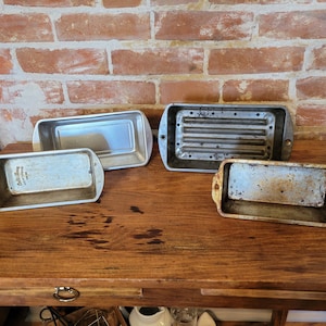 May include: Four silver loaf pans on a wooden table. The pans are different sizes and have varying degrees of wear and tear.