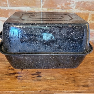 以下が含まれることがあります： 黒のエナメル製のローストパンで、斑点模様の仕上げになっています。パンには蓋が付いており、木製の表面の上に置かれています。