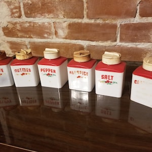 May include: Set of six vintage spice jars with red lids and white ceramic bodies. Each jar has a different spice label: Clove, Nutmeg, Pepper, Allspice, Salt, and Cinnamon.