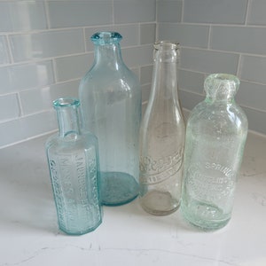 Puede incluir: Cuatro botellas de vidrio vintage con diferentes etiquetas y texto. Las botellas son transparentes y tienen un tono verdoso. Las botellas están sobre una superficie blanca.