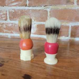 May include: Two vintage shaving brushes with white bristles and brown bands. The brush on the left has an orange base and the brush on the right has a red base. Both brushes have white handles.