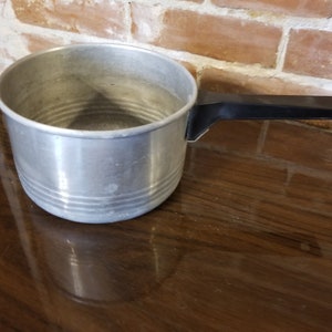 Pode incluir: Uma panela de alumínio prateada com uma alça de plástico preto. A panela tem uma aparência ligeiramente desgastada e está sentada em uma superfície de madeira marrom escura.