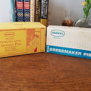 Puede incluir: Dos cajas de alfileres de costura Scovill. La caja amarilla es para alfileres de ropa de acero inoxidable y dice "Rustproof, Stiff Wire, Sharp Points". La caja blanca es para alfileres de modista de latón.