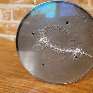 Pu&ograve; includere: Primo piano del fondo di un contenitore in metallo nero con la scritta "Factory No. 1" e "Dist. Ohio" stampata su di esso.