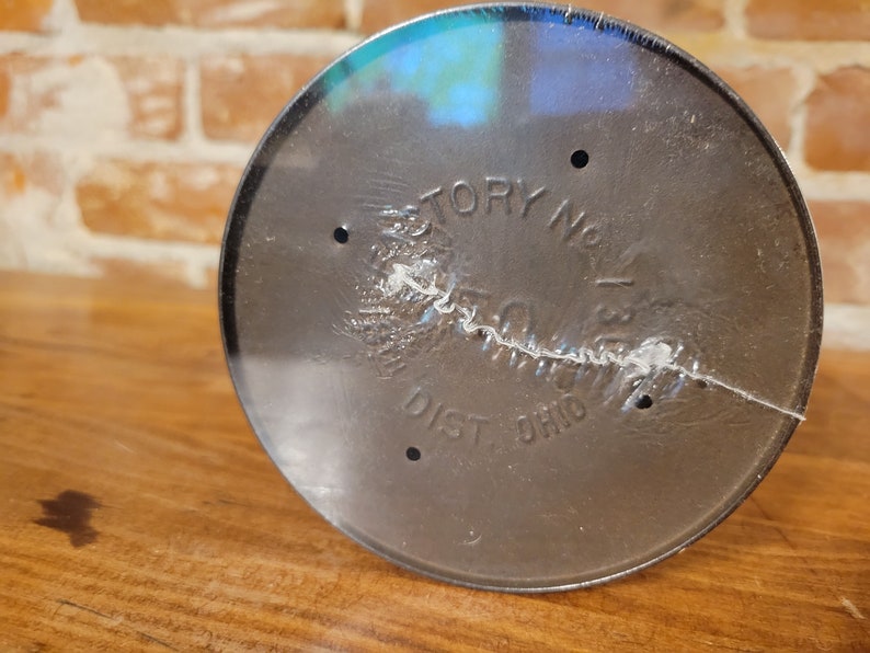 Pu&ograve; includere: Primo piano del fondo di un contenitore in metallo nero con la scritta "Factory No. 1" e "Dist. Ohio" stampata su di esso.