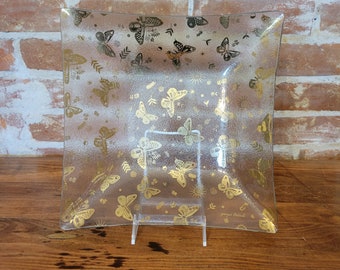 May include: A clear glass serving dish with a gold butterfly pattern. The dish has a slightly curved edge and is decorated with a variety of gold butterflies, leaves, and stars. The text "Joannes Michael" is printed on the bottom of the dish.