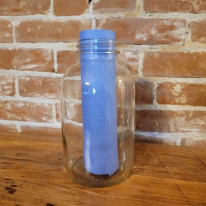 Puede incluir: Un tarro de vidrio transparente con un papel azul enrollado en su interior. El tarro está sobre una superficie de madera frente a una pared de ladrillo.