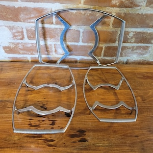 Puede incluir: Tres rejillas de enfriamiento de pastel de metal plateado. La rejilla más grande es de forma ovalada y las dos rejillas más pequeñas son rectangulares. Las rejillas están sobre una superficie de madera.