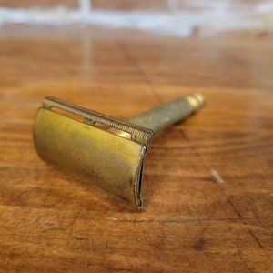 Puede incluir: Una maquinilla de afeitar de seguridad vintage de color dorado con una cabeza curva y un mango texturizado.