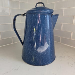 May include: A blue speckled enamelware coffee pot with a lid and handle. The pot has a spout for pouring and is sitting on a white countertop. The pot is a classic kitchenware item.