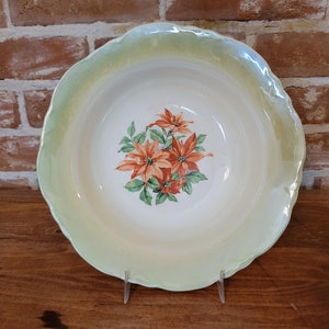 以下が含まれることがあります： 緑色の縁取りとオレンジ色のポインセチアの花の模様が施された白い陶器のボウル。ボウルは波打った縁をしています。