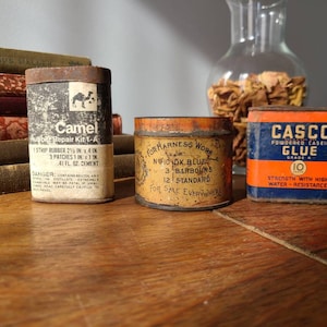 May include: Three vintage metal tins with labels. The first tin is orange and black and says "Camel Rubber Repair Kit 1-A". The second tin is brown and says "For Harness Work N10 DK. Blue 3 Barbours 12 Standard For Sale Everywhere". The third tin is blue and orange and says "Casco Powdered Casein Glue Grade A 10 Strength with High Water-Resistance".