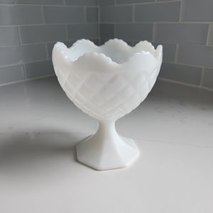 May include: White milk glass pedestal bowl with a scalloped rim and a geometric pattern. The bowl is on a white surface.