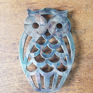 Puede incluir: Salvamanteles de plata con forma de búho con un diseño recortado. El búho tiene grandes ojos redondos y un cuerpo festoneado.
