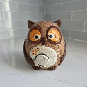 May include: A ceramic owl ornament in shades of brown and tan, featuring large orange eyes and a white hat adorned with orange flowers. The owl has a rounded body and sits upright.