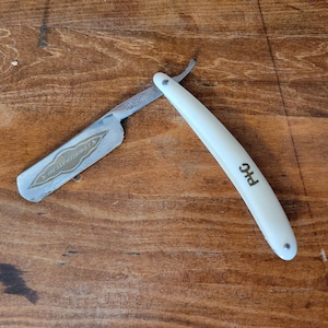 May include: A white straight razor with a gold-colored handle and a silver blade. The blade has the text "GROSS-SCHLEIFMÜHLE" and "IG 124" on it. The handle has the text "P+G" on it.