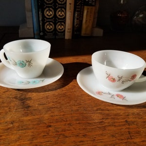 May include: Two vintage teacups and saucers with floral designs. The teacup on the left has blue flowers and the teacup on the right has pink flowers.