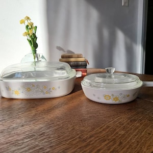 May include: Two white casserole dishes with floral designs in yellow and blue. The larger dish has a glass lid. Both dishes are on a wooden table.