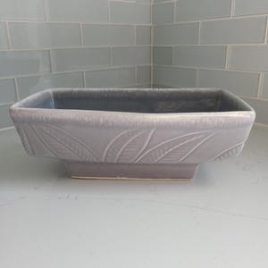 May include: A light grey ceramic planter with a rectangular shape and a leaf design. The planter has a slightly raised rim and a flat base.