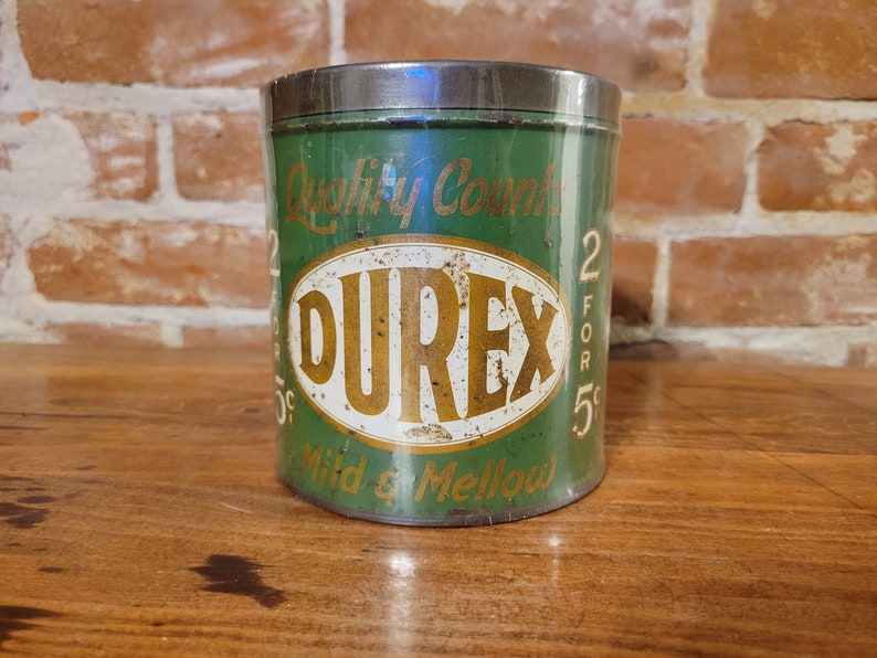 Pu&ograve; includere: Una scatola di metallo verde vintage con un'etichetta ovale bianca su cui &egrave; scritto "Durex" in lettere dorate. La scatola ha il testo "Quality Counts 2 for 5c Mild & Mellow" stampato su di essa.