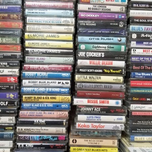 Może przedstawiać: Ściana kaset magnetofonowych z muzyką bluesową, w tym artystów takich jak Muddy Waters, Howlin' Wolf, Jimmy Reed, B.B. King i Otis Redding. Kasetki są ułożone w rzędach i kolumnach, a tytuły i nazwiska artystów są widoczne na grzbietach.