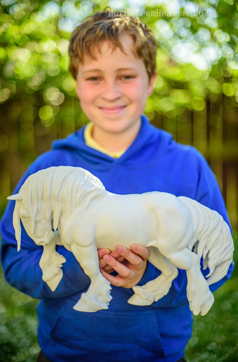 May include: A white horse figurine with a flowing mane and tail is held in front of a blurred green background. The horse is the focal point, with detailed sculpting. The figurine is held by a person wearing a blue hoodie.