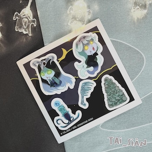 Peut inclure: Une planche d'autocollants avec des illustrations de personnages effrayants. Les dessins incluent un personnage cornu, une figure de sirène, un peigne, un tas de pierres et une bougie. La planche a une bordure blanche et est sur fond sombre.