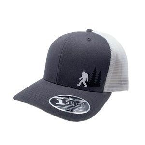 Può includere: Un cappellino da baseball grigio Flexfit 110 con retro in rete bianca. Il cappellino presenta una silhouette di Bigfoot e due alberi neri sul davanti. Il logo Flexfit 110 è sul davanti.